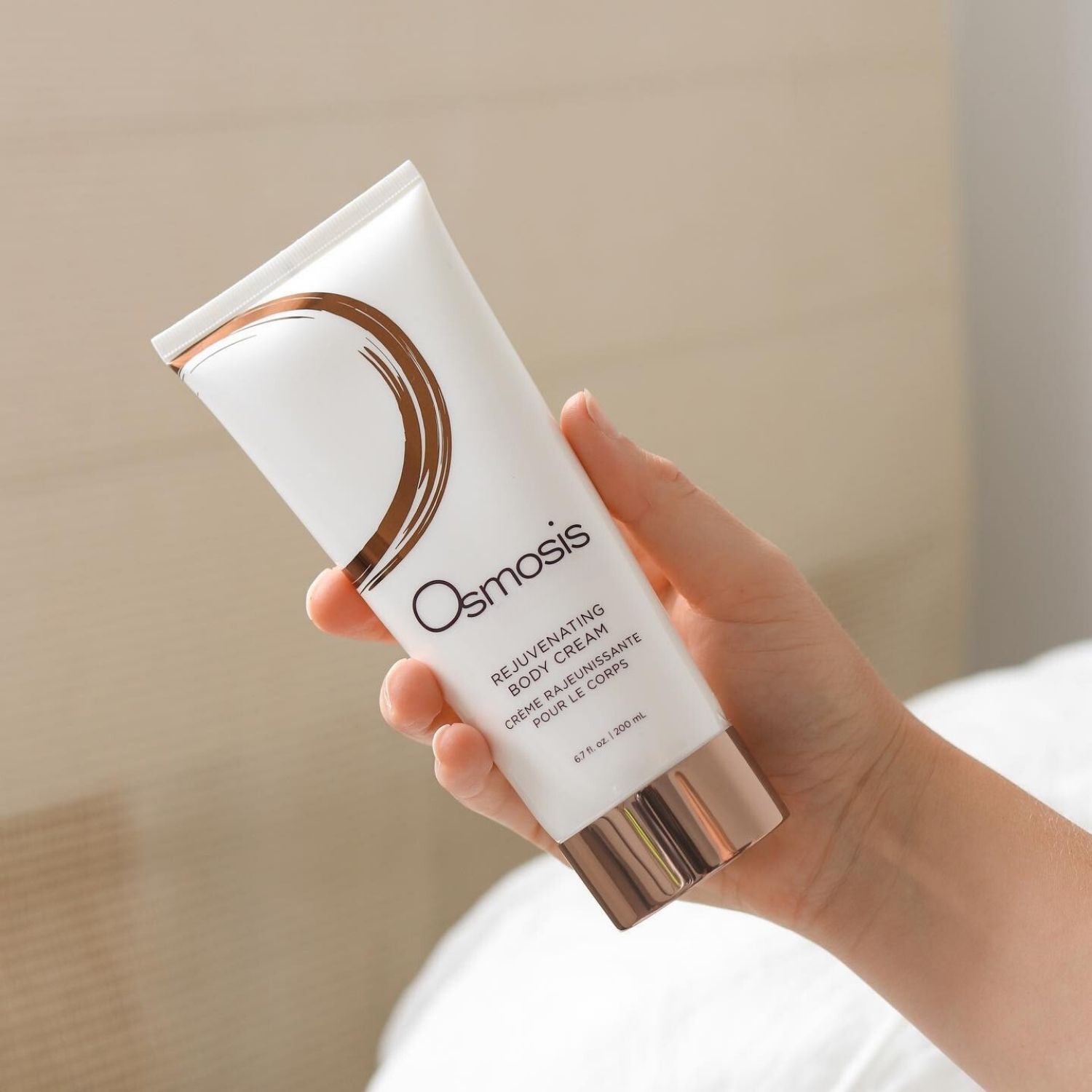 Hand holding a tube of Osmosis Rejuvenating Body Cream against a neutral background