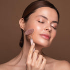 Woman using rose quartz skin roller on her face with tan background